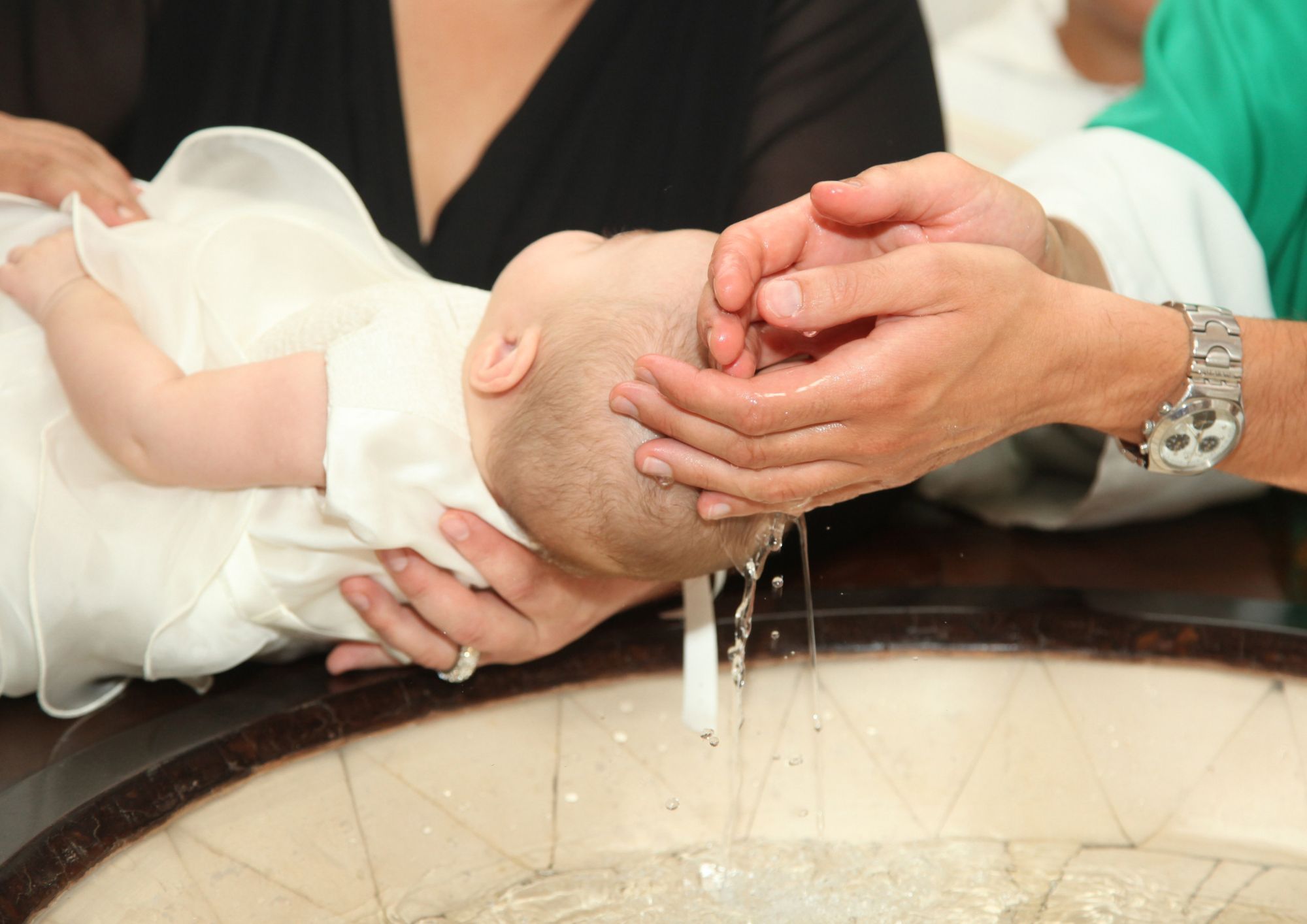 Billede af en baby som bliver døbt, hvor præsten hælder vand fra hænderne ud over babyens hoved.
